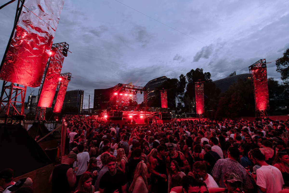 Large crowd dancing at Sidebyside Festival under red stage lighting at dusk with city skyline in background.